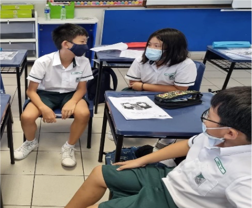 Three students in white uniforms and face masks sit at school desks in a classroom discussion.